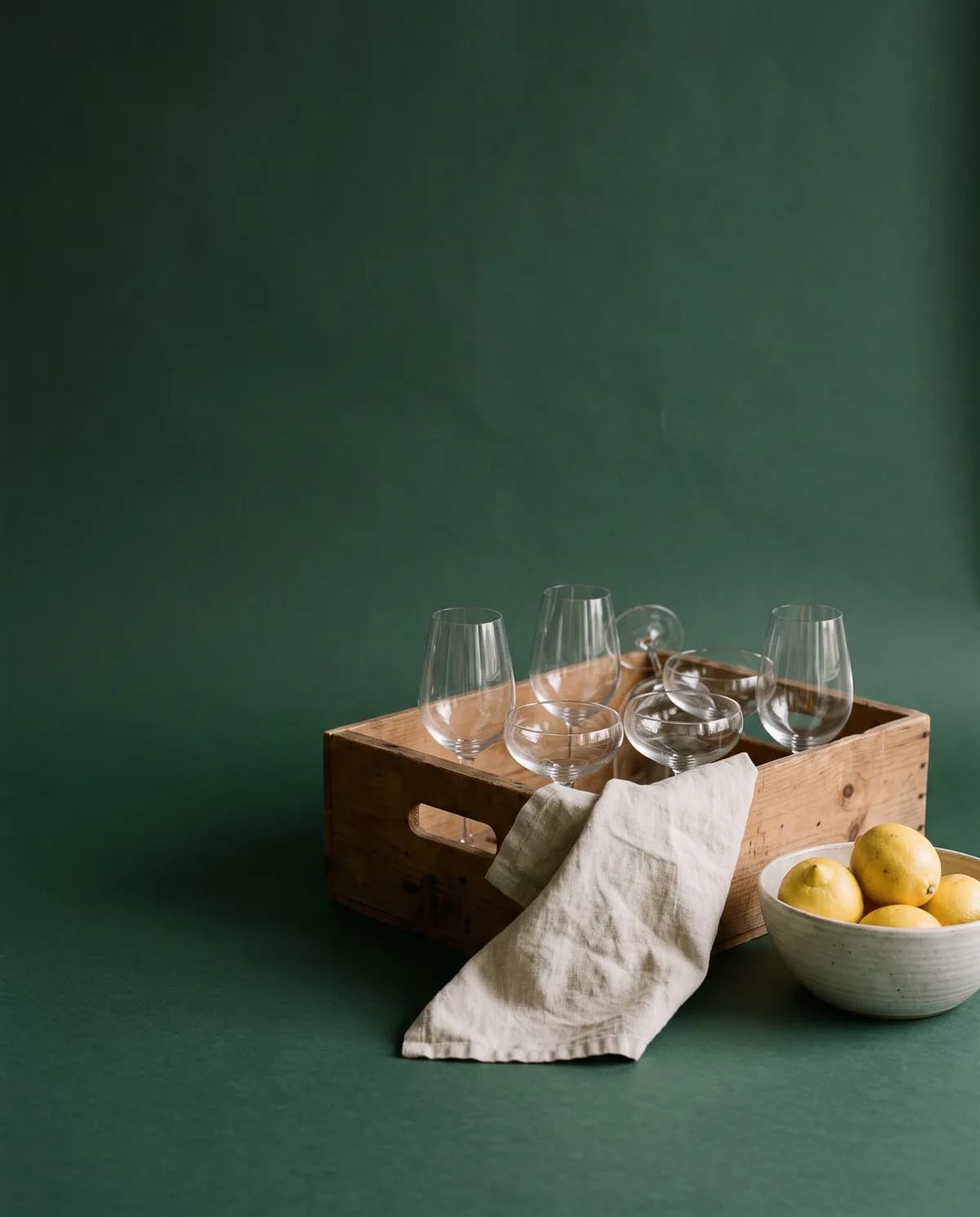 A wooden crate of wine glasses and coupes beside a linen cloth and a bowl of lemons